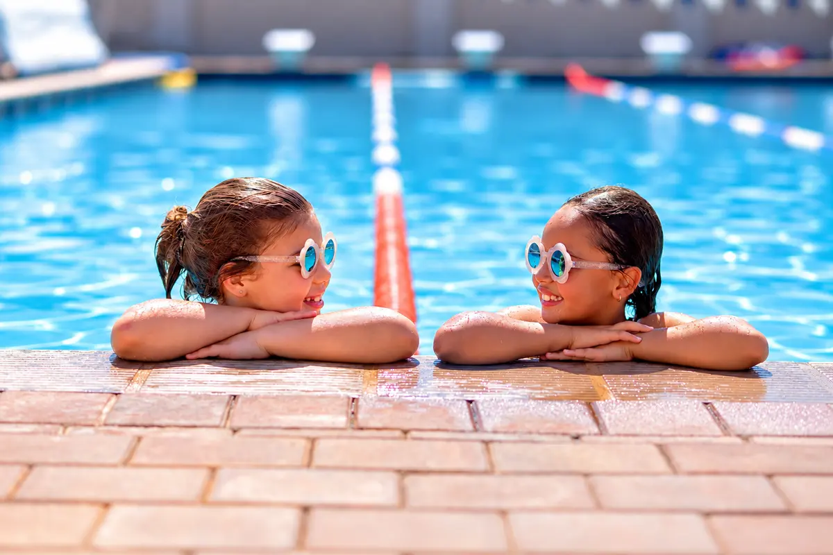 Two girls in goggles resting their chins on the edge of the pool, smiling