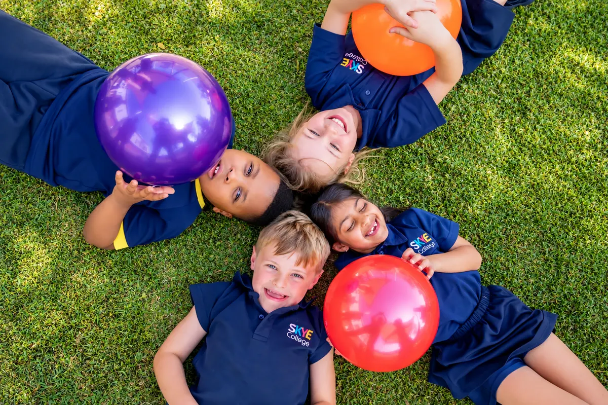 Five Skye College children lying in a circle on the grass with their heads together, laughing, holding bright balls