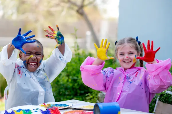 Two Junior Prep girls in painting smocks holding up paint-covered hands and laughing
