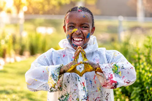 A child in a paint-spattered smock making a heart shape with painted hands