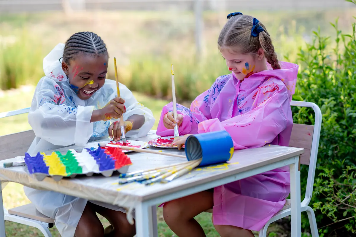 Two Prep girls in painting smocks laughing as they paint at an outdoor table